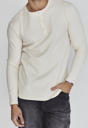 Mann trägt ein cremefarbenes Langarm-Henley-Shirt mit drei Knöpfen, schwarze Jeans und Armbänder am linken Handgelenk, steht mit der Hand in der Hosentasche.