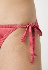 Rebrasti, koraljni donji dio bikinija s vezanjem sa strane. Materijal ima blagi sjaj, a tekstura djeluje mekano i fleksibilno.