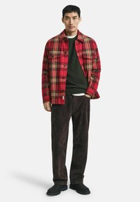 Mann in einer rot karierten Knopfjacke über einem dunkelgrünen Pullover, mit dunkelbraunen Hosen und schwarzen Schuhen, steht mit einer Hand in der Tasche.