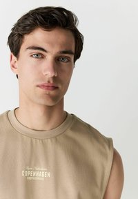 T-shirt sans manches beige avec un col rond et le texte "Copenhague" imprimé en couleur claire, confectionné dans un tissu doux au toucher lisse.