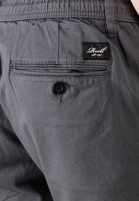 Pantalones cortos grises con una tela texturizada, que presentan un bolsillo trasero con un botón negro y una etiqueta cosida que dice "Reell" en letras cursivas blancas.
