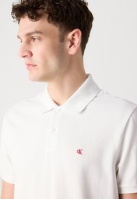 Jonge man met kort krullend haar, die een wit poloshirt met een rood "cK"-logo draagt, kijkt naar links tegen een effen lichte achtergrond.