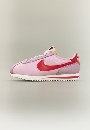 Lavskåret lyserød sneaker med rød Nike-swoosh, hvide snørebånd, rød hælkappe og hvid sål med rød stribe, vist fra siden på en neutral baggrund.