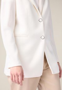Hvid skræddersyet blazer med to runde sølvknapper og en enkelt brystlomme, parret med lys beige bukser. Glat stoftekstur.