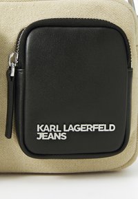 Beige fabric bag with a black leather zippered pocket. Features silver "KARL LAGERFELD JEANS" lettering on the leather accent.