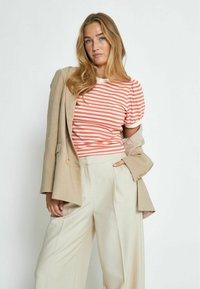 Maglietta a righe bianca e corallo con maniche a sbuffo, abbinata a pantaloni beige a gamba larga e un blazer beige chiaro. Trame lisce.