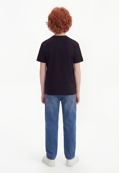 Παιδί με σγουρά κόκκινα μαλλιά, που φοράει μαύρο T-shirt, μπλε τζιν και λευκά αθλητικά παπούτσια, στέκεται με την πλάτη προς το μέρος μας σε λευκό, ομοιόμορφο φόντο.