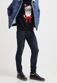 Granatowy sweter z grafiką Świętego Mikołaja, kurtka dżinsowa z ociepleniem z wełny, ciemne dżinsy oraz czarne buty z jasnobrązowymi podeszwami.