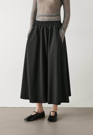 Person iført en lang sort nederdel med elastisk talje og lommer, kombineret med sorte Mary Jane-flade sko og en semi-gennemsigtig bluse med lange ærmer.