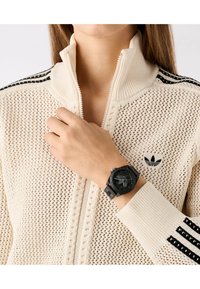 Montre noire avec cadran texturé et bracelet en caoutchouc portée sur une main contre une veste à zip en tricot de couleur crème avec des rayures noires.