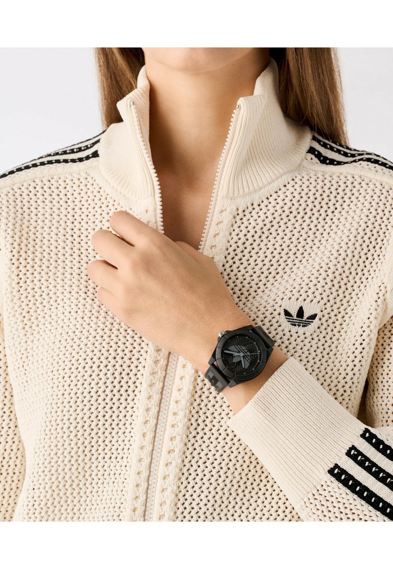 Montre noire avec cadran texturé et bracelet en caoutchouc portée sur une main contre une veste à zip en tricot de couleur crème avec des rayures noires.