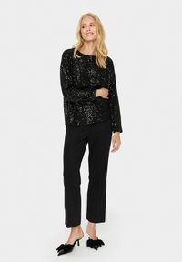 Top lungo con maniche nere in paillettes abbinato a pantaloni neri a gamba dritta e mules neri con tacco e dettagli a fiocco. Design semplice ed elegante.