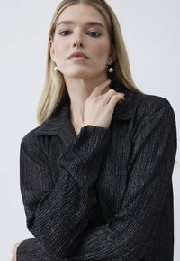 Maglione nero con texture, colletto e motivo a coste. Il modello indossa orecchini sferici argentati e tocca il collo, mettendo in mostra il tessuto.