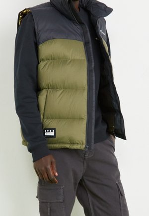 Mężczyzna ubrany w oliwkowo-zieloną i ciemnoszarą pikowaną kamizelkę na czarnej koszuli z długim rękawem, w ciemnoszarych spodniach typu cargo, stojący na białym tle.