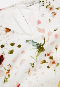 Weiße V-Ausschnitt-Bluse mit einem zarten Blumenmuster in den Farbtönen Rosa, Grün und Rot, mit dem Etikett Cellbes of Sweden.