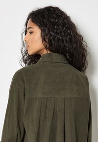 Olivgrünes Cordhemd mit Kragen, das ein plissiertes Rückendetail und eine strukturierte Oberfläche aufweist. Im lässigen Fit gestaltet.