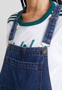 Lähellä olevan henkilön kuva, jolla on ylläsininen farkkuhaalarit messinkisillä soljilla valkoisen paidan päällä, jossa on vihreä kaulus ja osittainen teksti.