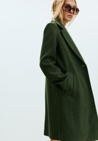 Zelený kabát ze směsi vlny s klopami, jednobarevným uzavíráním na jeden knoflík a bočními kapsami. Má uvolněný střih a měkkou strukturu.