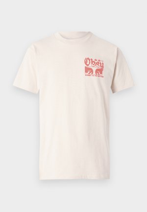 Kratkotrajna, svetlo bež bombažna majica s kratkimi rokavi, ki ima na sprednji strani rdečo grafiko z napisom "Obey" in "Submit to the rhythm".