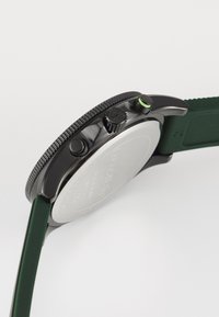 Montre chronographe noire avec un bracelet en silicone vert, un boîtier en acier inoxydable, une lunette texturée et trois boutons sur le côté.