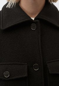 Cappotto di lana nera con un ampio colletto, due tasche frontali e bottoni neri. La texture è morbida e leggermente pelosa.