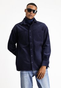 Camicia di velluto a coste navy con due tasche sul petto, chiusura con bottoni e vestibilità rilassata, abbinata a jeans di denim blu. Colletto piatto e maniche lunghe.
