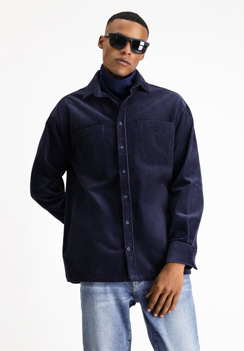 Camicia di velluto a coste navy con due tasche sul petto, chiusura con bottoni e vestibilità rilassata, abbinata a jeans di denim blu. Colletto piatto e maniche lunghe.