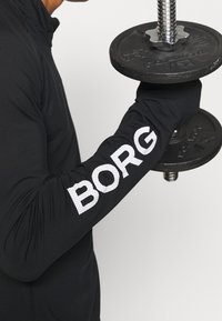 Schwarzes Langarmoberteil mit "BORG" in weißen, gepunkten Buchstaben auf dem Ärmel, der einen schwarzen Hantel mit einem silbernen Griff hält.
