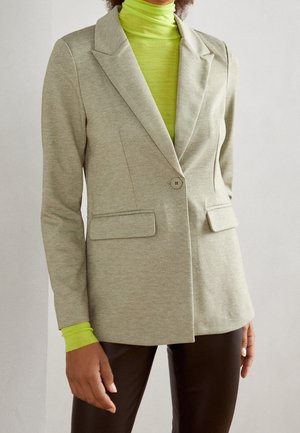 Vrouw draagt beige blazer met één knoop over limoengroene coltrui en donkerbruine broek, poserend tegen een effen lichte muur.