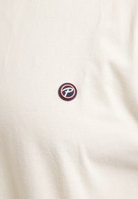 Krémové bavlněné tričko s jemnou texturou, které má kulatou vyšívanou nášivku se stylizovaným "P" v námořnické modré a červené akcenty.