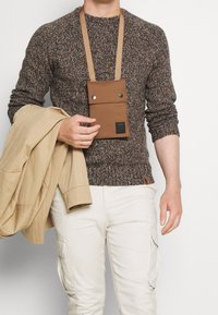 Braune Umhängetasche mit strukturierter Oberfläche, zwei Druckknöpfen und abnehmbarem Riemen; getragen von einer Person in einem braun gesprenkelten Pullover und einer beige Jacke.