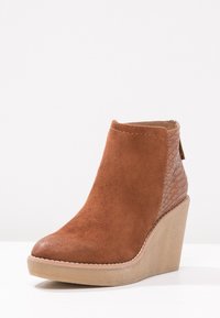 Botín de tobillo en ante marrón con un tacón texturizado, punta redondeada y un costado liso. Presenta un diseño con costuras mínimas y una cremallera discreta.