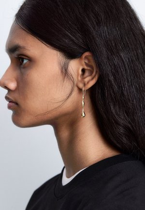 Gold drop earrings feature an elongated, organic shape with a smooth surface. The model has dark hair and wears a black shirt with a white collar.