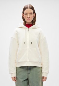 Cremefarbener Reißverschluss-Hoodie mit großer Kapuze, geräumigen Fronttaschen und gerippten Bündchen. Über einem roten Rollkragenpullover getragen, mit schlichten Designelementen.