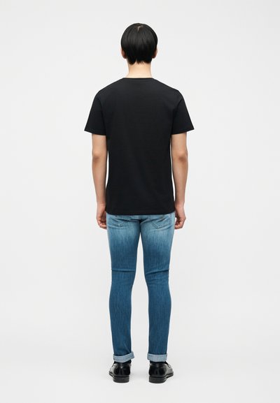 Άτομο που στέκεται με την πλάτη, φοράει μαύρο απλό μπλουζάκι, ξεθωριασμένα μπλε τζιν γυρισμένα στις μανσέτες και μαύρα παπούτσια φόρμας.