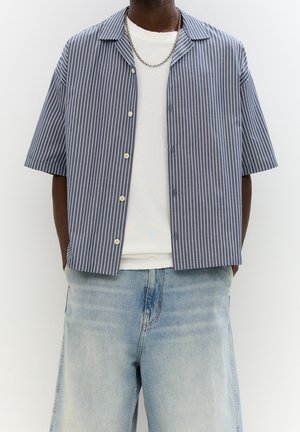 Man draagt een blauw-wit verticaal gestreept overhemd met korte mouwen over een wit T-shirt en lichtblauwe spijkerbroek.