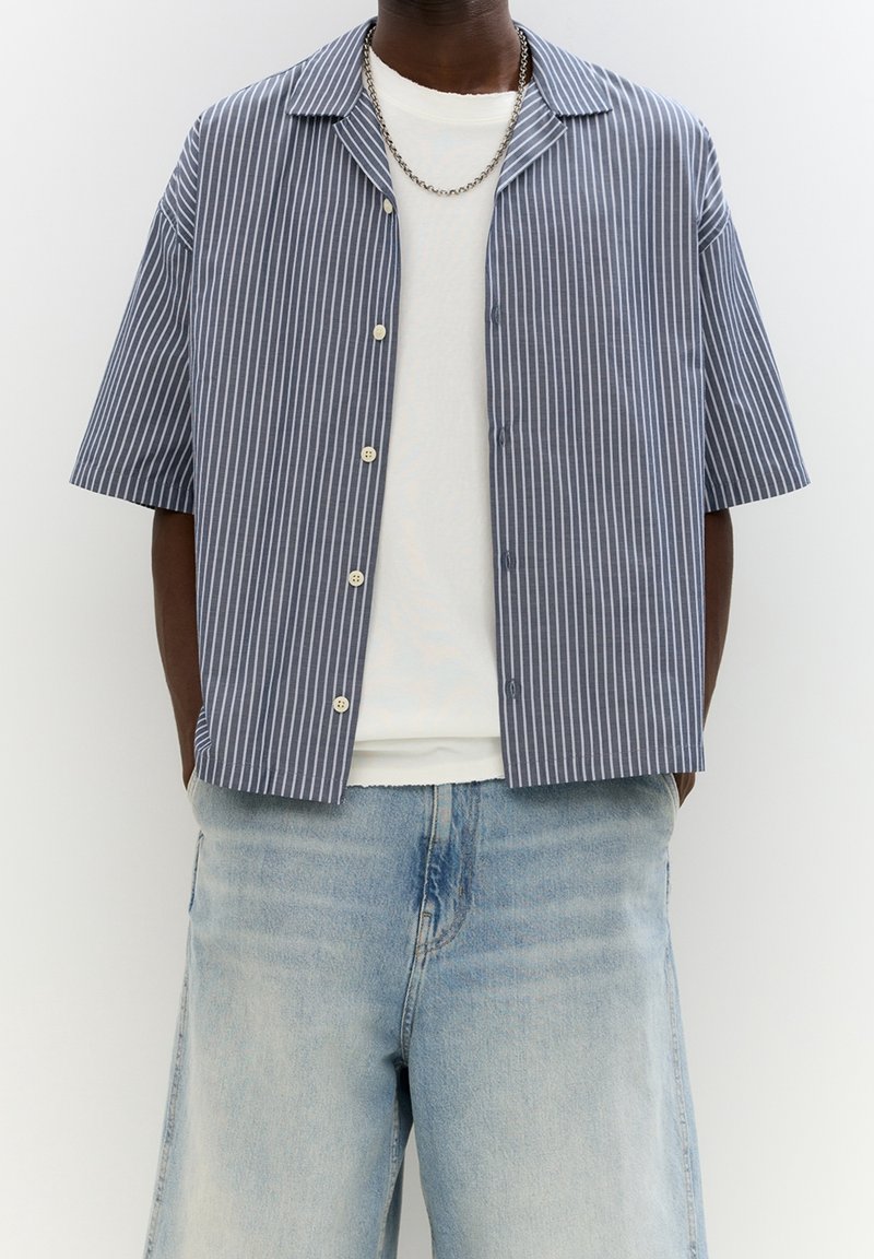Homme portant une chemise à manches courtes rayée verticalement bleu et blanc par-dessus un t-shirt blanc avec un jean bleu clair.