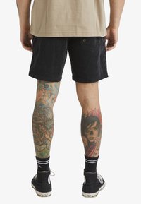 Shorts en velours côtelé noirs avec une coupe décontractée, tatouages détaillés sur les deux jambes, et baskets noires avec des accents blancs. Look décontracté et contemporain.