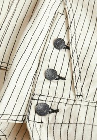 Chemise à boutons, en tissu crème avec des rayures verticales noires ; quatre boutons-pression noirs et détails de couture contrastants.