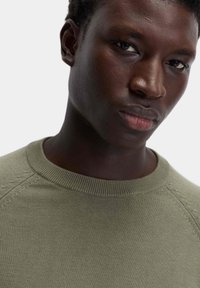 Maglione a maglia verde oliva con scollatura rotonda, tessuto testurizzato e cuciture dettagliate lungo le spalle. Primo piano della parte superiore del torso.