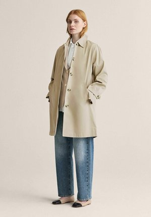 Kvinde iført beige trenchcoat over tan vest og hvid skjorte, kombineret med blå jeans og beige flade sko med sorte tåkapper, stående mod en ensfarvet baggrund.