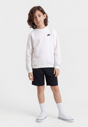 Enfant aux cheveux mi-longs portant un sweat-shirt Nike blanc, un short noir, des chaussettes blanches et des baskets blanches, debout devant un fond uni.