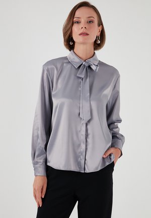 Silky, light gray blouse with a collar and bow tie detail, long sleeves, and button-down front, paired with dark trousers.