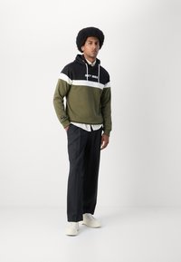 Olivengrønn, svart og hvit hettegenser med grafisk logo; kombinert med svarte bukser og hvite sneakers. Modellen står oppreist.