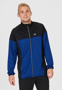 Blå og sort zip-up jakke lavet af letvægtsstof, med lange ærmer, høj krave og et logo på venstre bryst.
