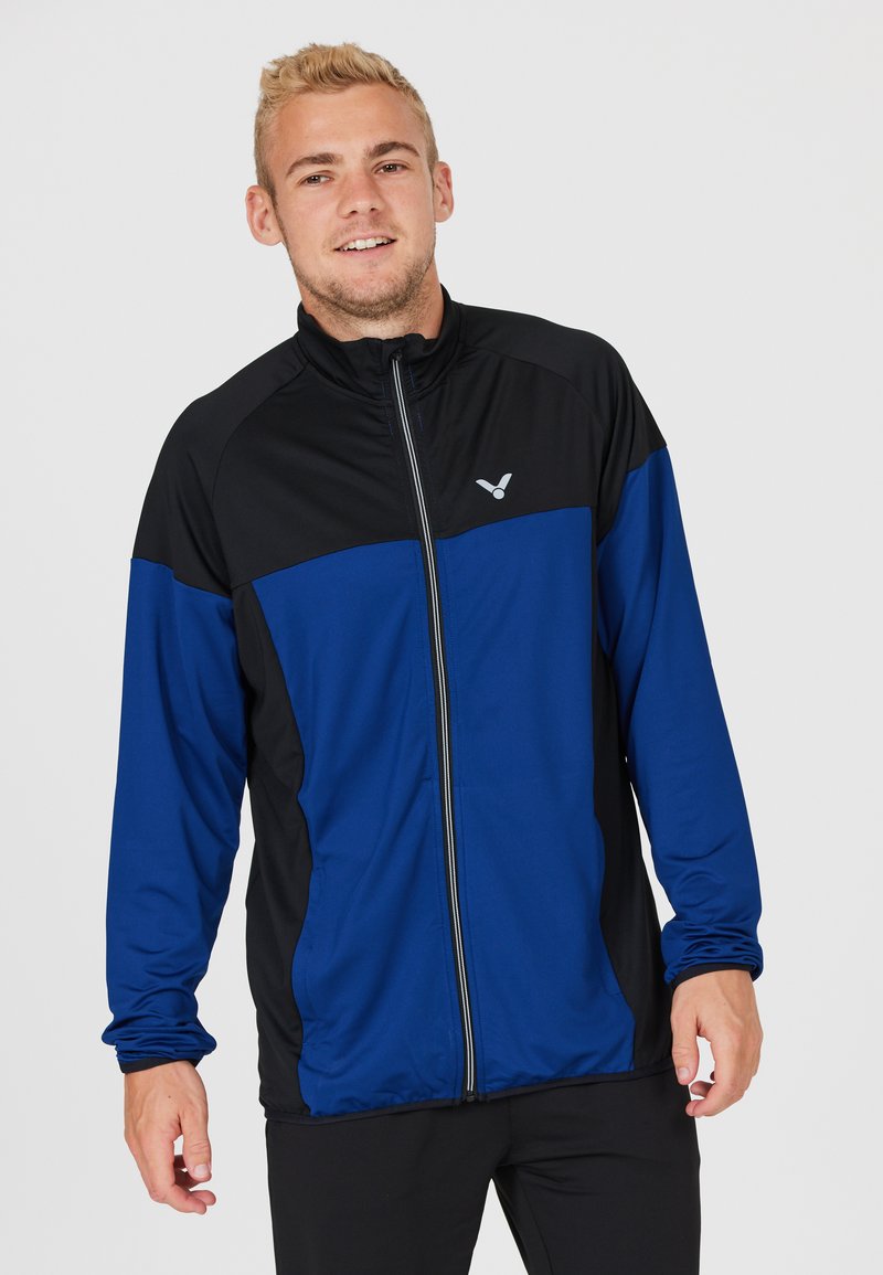 Blå og sort zip-up jakke lavet af letvægtsstof, med lange ærmer, høj krave og et logo på venstre bryst.