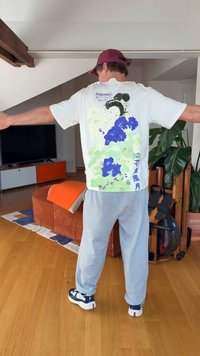 Άνδρας που φοράει λευκό t-shirt με καλλιτεχνικό σχέδιο με λουλούδια και γυναίκα, γκρι φόρμες και κόκκινο καπέλο, στέκεται με ανοιχτά τα χέρια σε εσωτερικό χώρο.