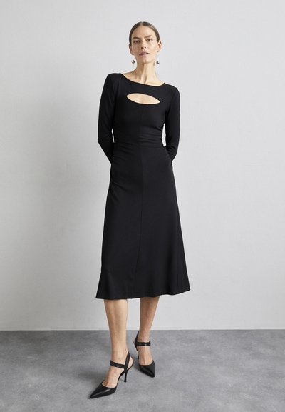 Μαύρο φόρεμα με μακριά μανίκια και λεπτομέρεια ανοίγματος μπροστά, εφαρμοστό σουρωτό και φαρδιά βάση. Φορεμένο με μαύρες γόβες με μυτερή μύτη και λουριά στον αστράγαλο.