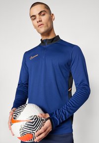 Kék, hosszú ujjú sportfelső negyedzippes nyakkal és narancssárga részletekkel, fekete-fehér mintázatú focilabdával párosítva.
