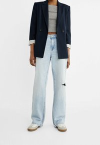 Blazer navy con due bottoni e maniche arrotolate, abbinato a jeans larghi chiari strappati e sneakers bianche con suole marroni.